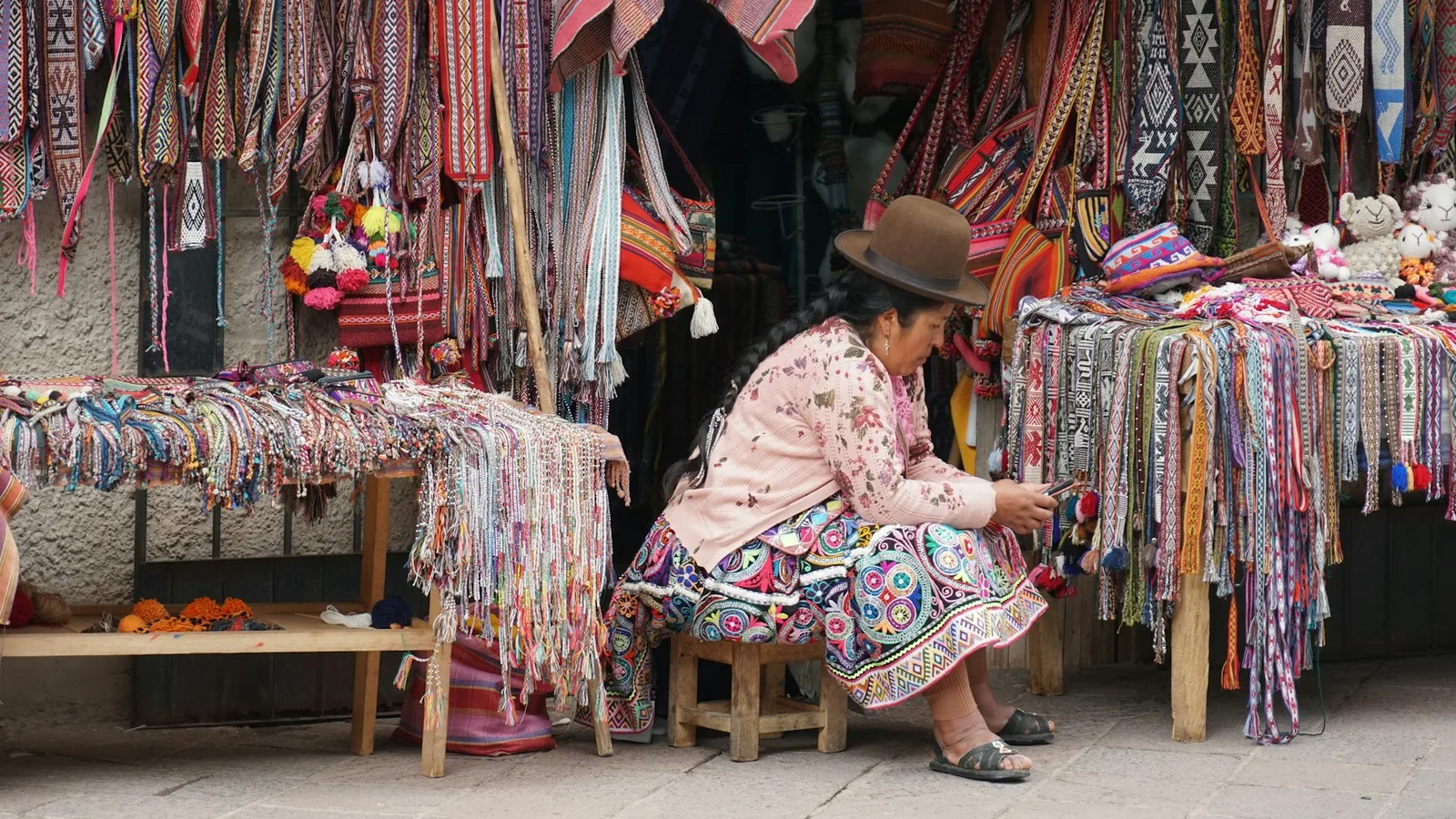 A craft stall in Pisac, Peru, with woven bands, bags and soft toys on display and a vendor seated on a stool